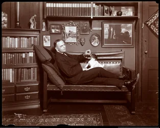 Will R. Barnes allongé sur un canapé avec un chien dans son studio bibliothèque, 1903